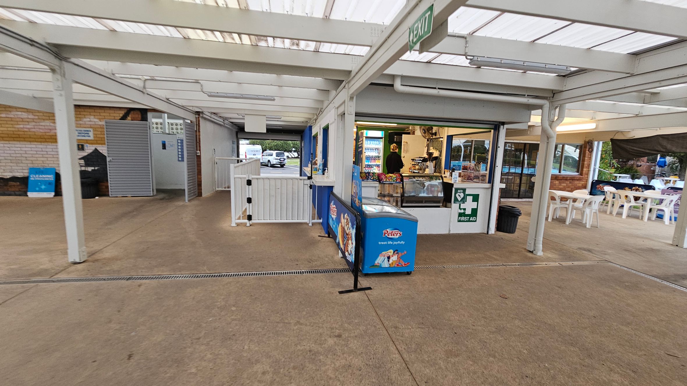 A covered outdoor area with a concrete floor. On the right, there's a small shop with a counter displaying snacks and a fridge with drinks. A first aid sign is visible. To the left, a grey metal door and a blue cleaning sign are against a brick wall. In the background, white plastic tables and chairs are set up. The ceiling is made of translucent panels, and an "EXIT" sign hangs above.
