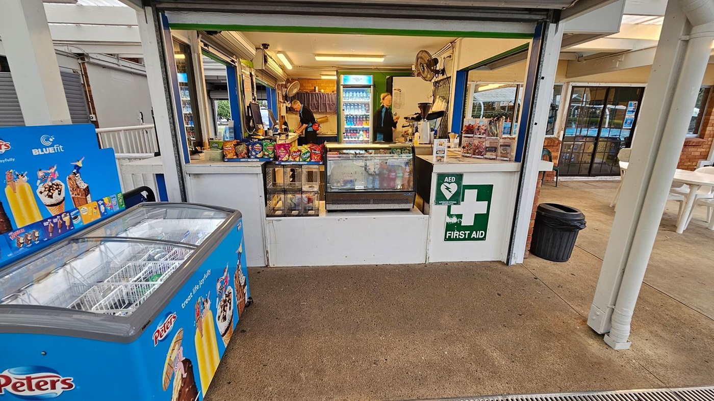 A small kiosk with a counter displaying snacks and a glass-fronted fridge filled with drinks. Two people are inside, one near the counter and another by a fridge in the back. To the left, there's a blue ice cream freezer with branding. A first aid sign is visible on the right. The floor is concrete, and there are white plastic chairs and tables in the background near a glass door. A black rubbish bin is positioned to the right of the kiosk.