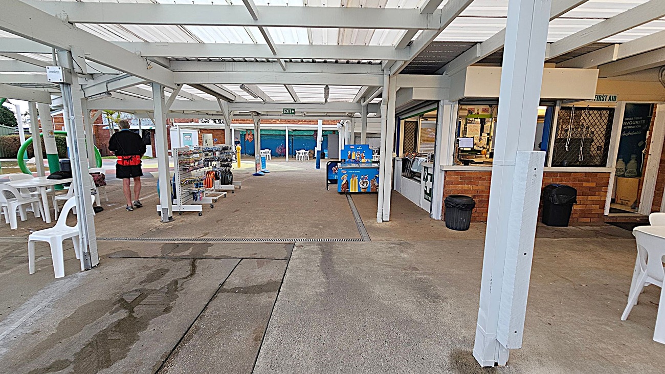 A covered outdoor area with a concrete floor. On the left, there are white plastic chairs and tables, and a person walking away holding an umbrella. In the centre, there are display racks with various items. On the right, a brick kiosk with a serving window and a sign for first aid. The ceiling is made of translucent panels supported by white beams. In the background, there are more tables and chairs, and a mural on the wall.