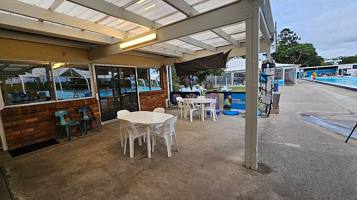 A covered outdoor area with a transparent roof and brick walls. On the left, there are two green plastic chairs against the wall. In the centre, white plastic tables and chairs are arranged on a concrete floor. To the right, a sign reads "POOL CLOSED" near a swimming pool. A lifeguard in orange is visible by the pool, and trees are in the background. The area is well-lit with fluorescent lights.