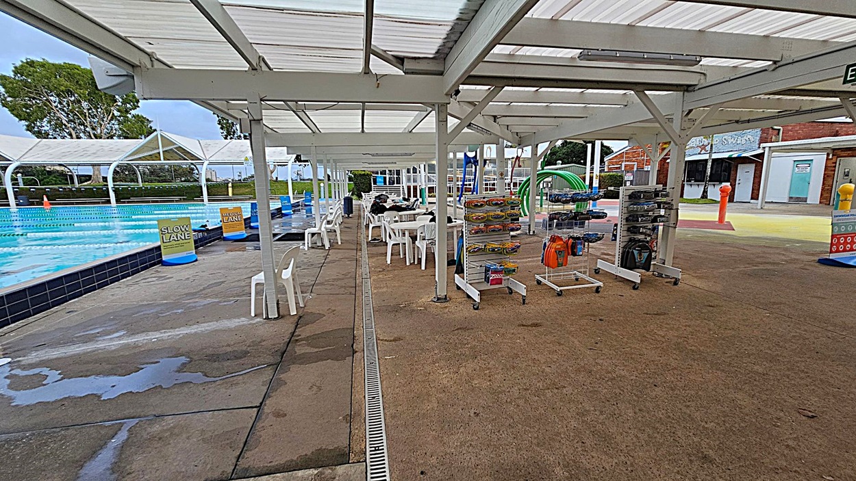 Outdoor swimming pool area with a covered walkway. On the left, a pool with lane markers and signs for "Slow Lane" and "Medium Lane". White plastic chairs and tables are under the shelter. On the right, racks with swimming gear and accessories. The ground is concrete, and the ceiling is a white, slatted structure. In the background, there are brick buildings and a grassy area with trees.