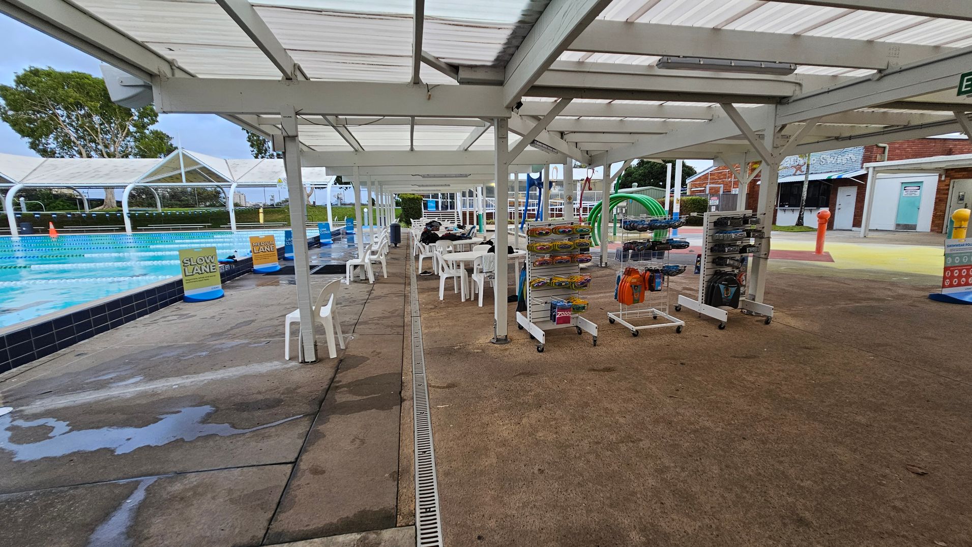 Outdoor swimming pool area with a covered walkway. On the left, a pool with lane markers and signs for "Slow Lane" and "Medium Lane". White plastic chairs and tables are under the shelter. On the right, racks with swimming gear and accessories. The ground is concrete, and the ceiling is a white metal structure. In the background, a brick building with doors and a sign. Greenery and trees are visible beyond the pool.