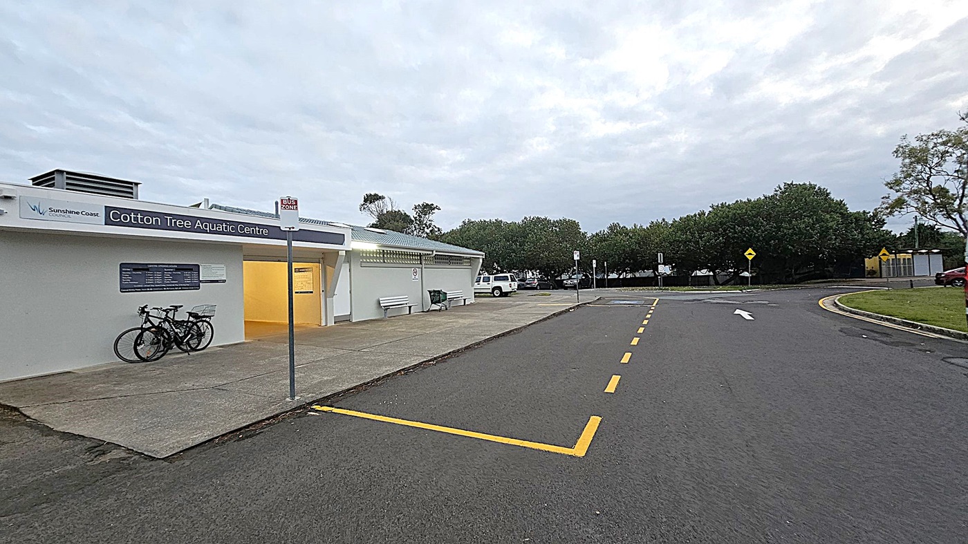A car park with two vehicles in designated disabled parking spaces. On the left, a black Mazda with a visible licence plate. To the right, a grey SUV. Behind the cars, a low building with a sign reading "Clifton Hill Aquatic Centre" is partially visible. Surrounding the area are green bushes and trees. A streetlamp is lit near the left side, and the ground is covered with dark asphalt.