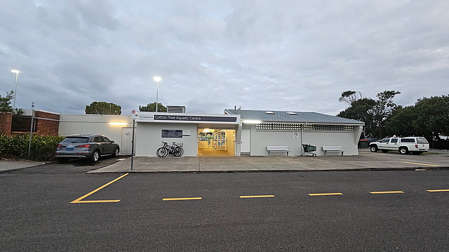 A single-storey building with a sign reading "Cotton Tree Aquatic Centre" above the entrance. Two bicycles are parked to the left of the entrance. A grey car is parked on the left side of the building, near some greenery. On the right, there are two white benches and a green shopping trolley. A white vehicle is parked further to the right. The building has a light grey exterior and a blue roof. The foreground shows a dark asphalt road with yellow markings.