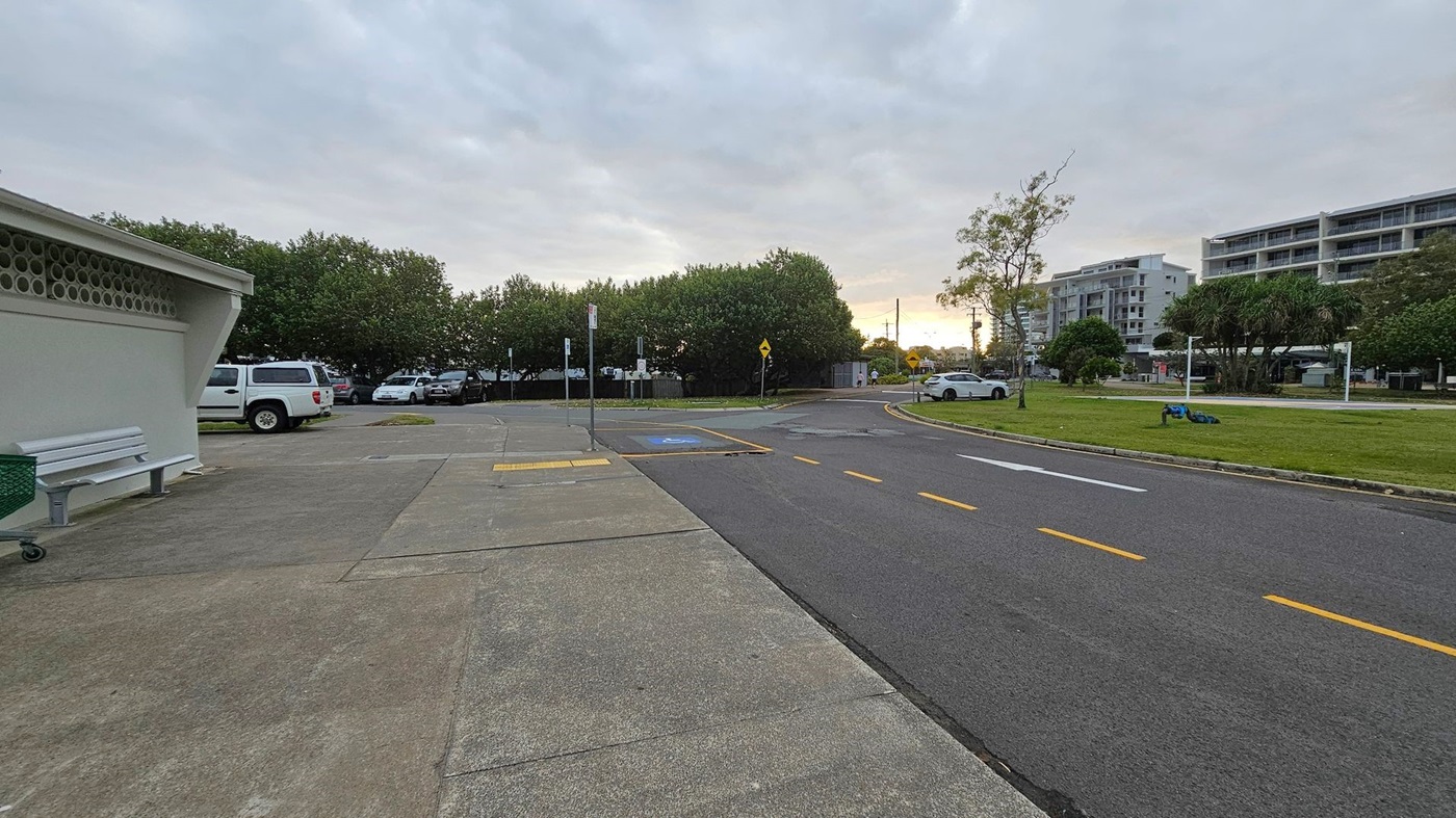 A paved area with a bench and a green bin on the left, next to a building with a perforated wall. Several parked cars are visible in the background, partially obscured by trees. On the right, a road with yellow dashed lines runs alongside a grassy area with a few trees and a white car. In the distance, there are multi-storey buildings. The sky is overcast, and the scene appears to be at dusk or dawn.