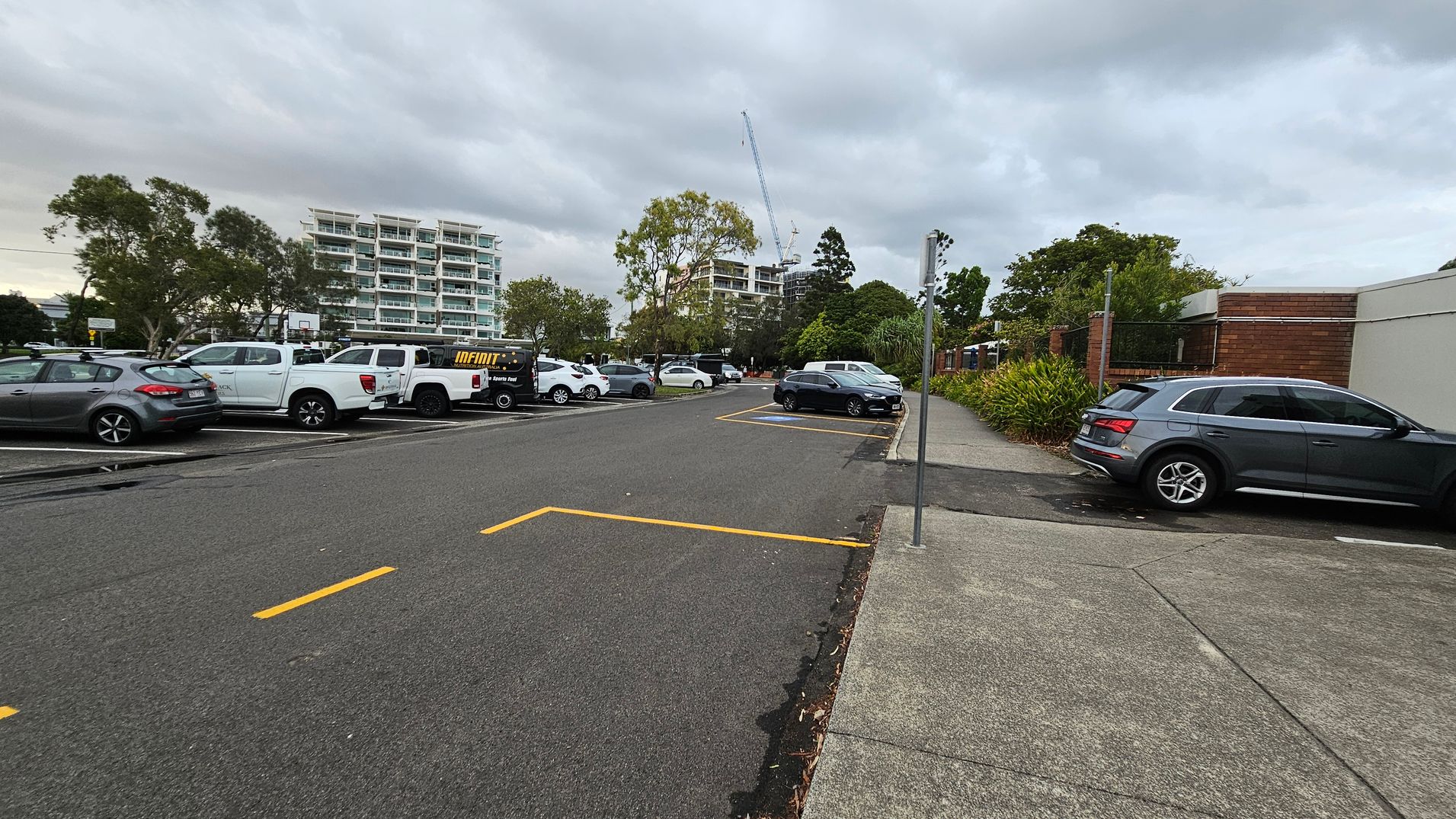 A car park with several vehicles on the left, including a white pickup truck and a black van. In the background, there are tall buildings and a construction crane. On the right, a grey car is parked next to a brick wall with greenery. The ground is asphalt with yellow parking lines, and the pavement on the right is light grey. Trees are scattered throughout the area.