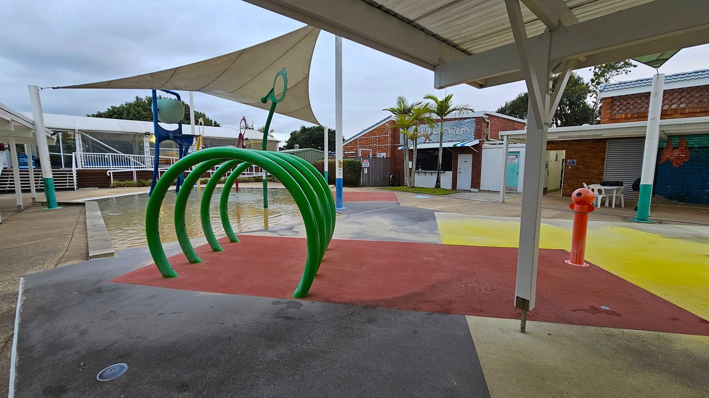 A playground with a splash pad area featuring a series of green arches on the left. The ground is covered with colourful rubber surfaces in red, yellow, and green. In the background, there are brick buildings with blue roofs and a few palm trees. A white shade sail is overhead, and a red water feature stands on the right. The area is surrounded by white railings and a small pool of water.