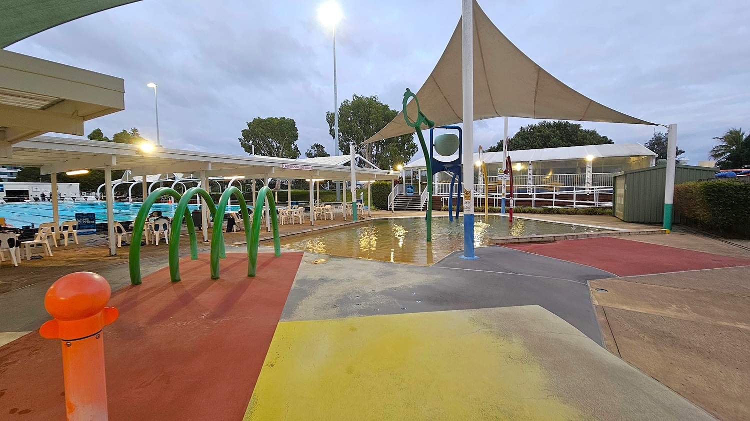 Outdoor swimming pool area with a splash pad in the foreground. The splash pad has green arches and a blue and white play structure. The ground is painted in various colours, including red, yellow, and grey. To the left, there are covered seating areas with white chairs. In the background, a large pool is visible, along with trees and a white canopy. The area is lit by tall lights, and the sky is overcast.