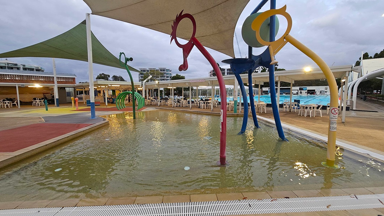 A children's splash pad with shallow water features colourful, curved structures resembling abstract animals. On the left, a green structure with loops stands near a red one. On the right, a blue and yellow structure is visible. In the background, there are white chairs and tables under a covered area, with a swimming pool beyond. The ground is a mix of red, yellow, and grey surfaces, and a green shade sail is overhead.