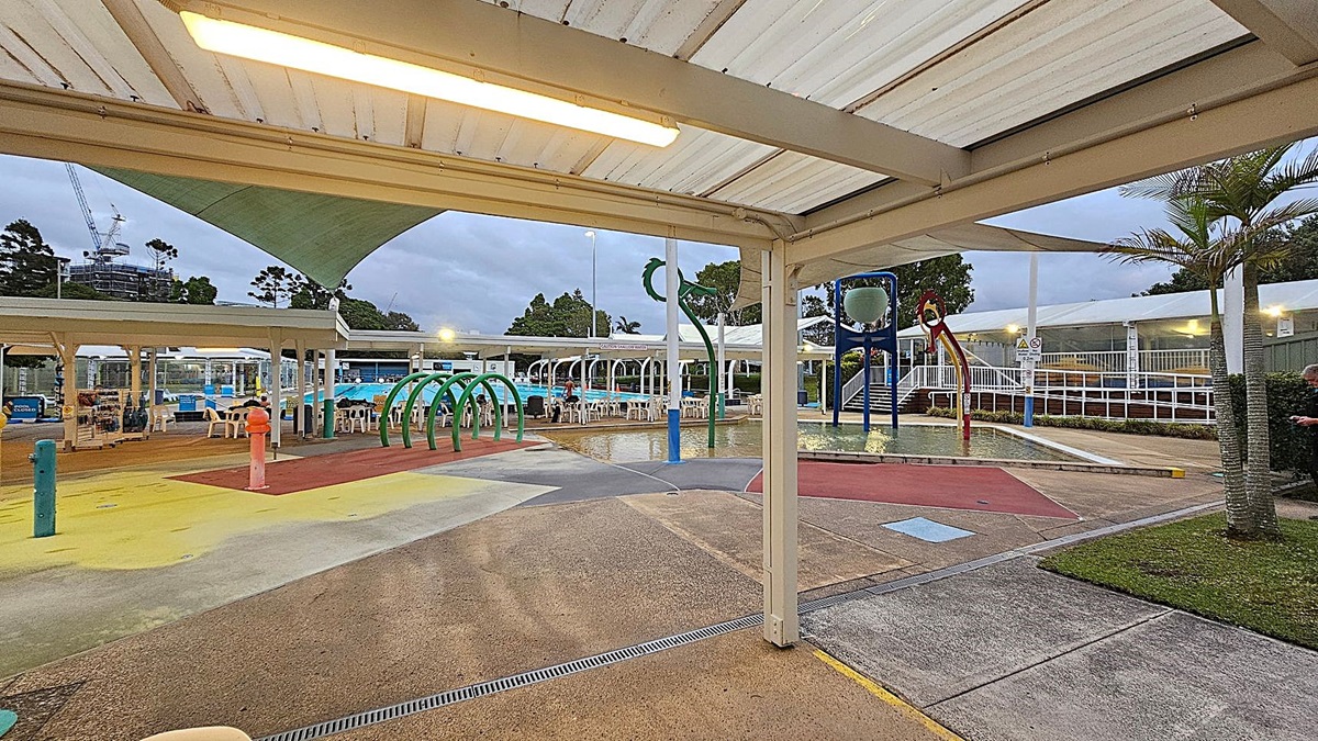 Outdoor water play area with colourful splash pads and water features. On the left, there are green arches and a red water spout. In the centre, a large bucket and slide are visible. The background has a swimming pool with white chairs and tables under a canopy. Trees and a construction crane are visible in the distance. The ground is a mix of concrete and coloured surfaces, and a palm tree is on the right. The ceiling is a white, slatted structure.