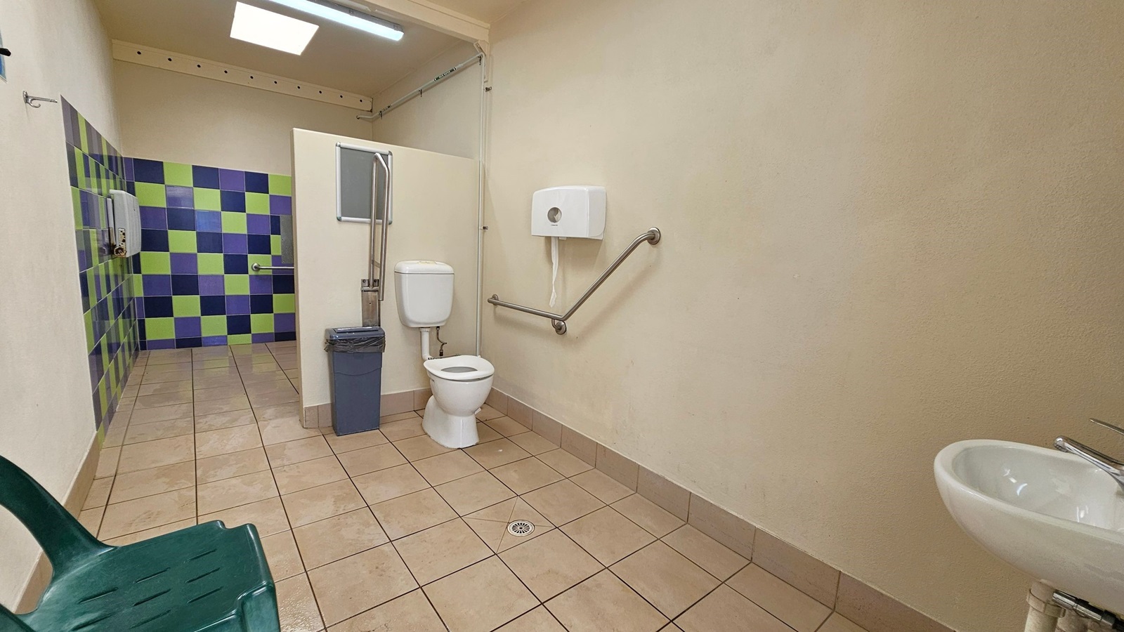 A public restroom with a tiled floor. On the right, there's a white toilet with a grab bar and a toilet paper dispenser above it. A small sink with a tap is also on the right. On the left, a green plastic chair is visible. The back wall features a colourful pattern of green and blue tiles. A waste bin is positioned near the toilet. The ceiling has a fluorescent light and a skylight.