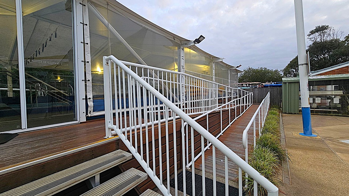 A set of metal and wooden steps leads up to a glass entrance with a "Pool Closed" sign. The entrance is flanked by white railings. On the left, there is a dense green hedge with red plants, while on the right, a wooden panelled wall and more railings are visible. The ground is a mix of textured black and concrete surfaces. Inside, a pool area is partially visible through the glass.