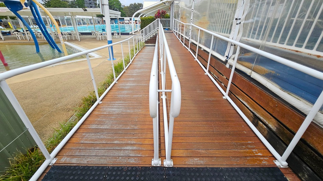 A wooden ramp with white metal railings runs straight ahead. On the left, there's a shallow water play area with colourful water features and a pool surrounded by white chairs and tables. On the right, a transparent barrier with a white frame lines the ramp. The ground is a mix of wooden planks and textured surfaces, with some grass visible on the left. In the background, there are trees and more poolside seating.