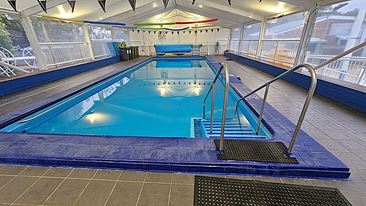 An indoor swimming pool with clear blue water, surrounded by a tiled floor. On the right, a metal handrail leads into the pool. The ceiling is supported by a white structure with triangular flags hanging across. The walls are transparent, allowing a view of the outside. At the far end, there are pool cleaning equipment and colourful pool noodles. The floor is grey with black non-slip mats near the pool's edge.