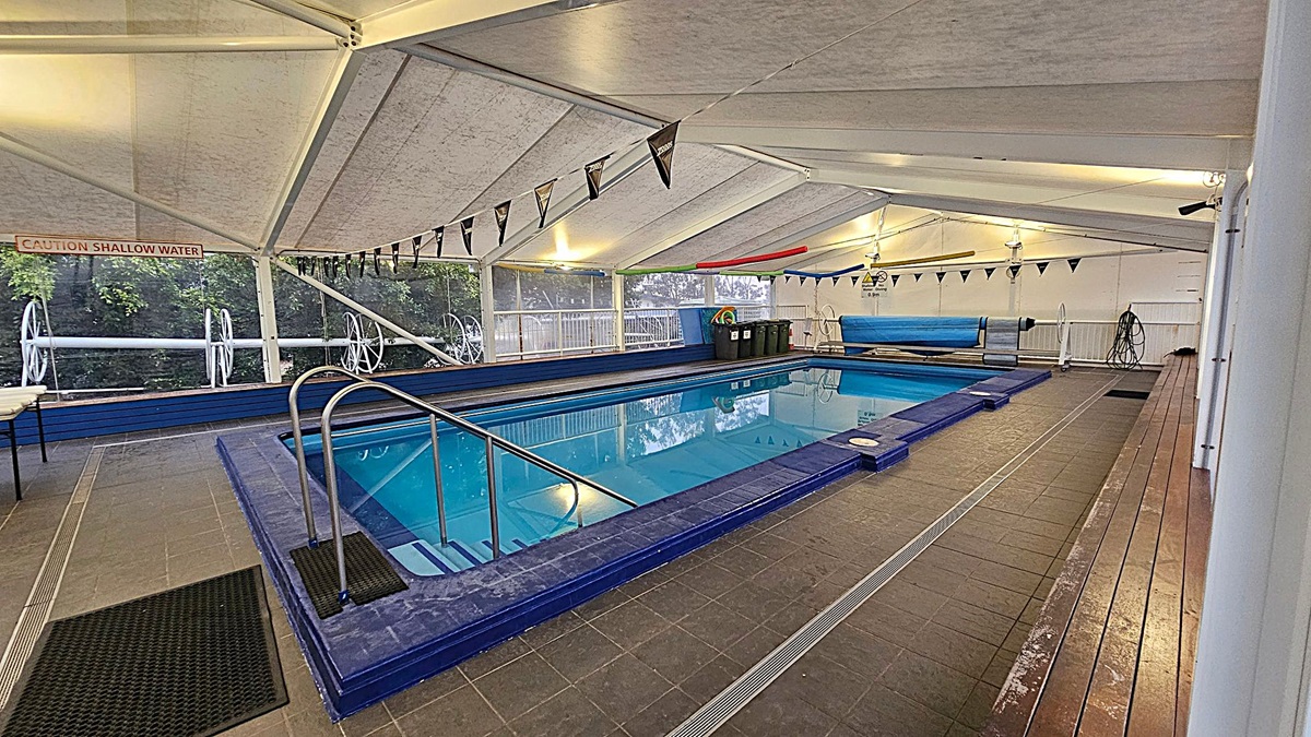 Indoor swimming pool with a blue-tiled edge and metal handrails on the left. The pool is surrounded by a tiled floor with black mats. A "Caution Shallow Water" sign is visible on the left wall. The ceiling is white with triangular flags hanging across. On the right, there are pool covers and equipment, including a hose. Large windows on the left and back walls reveal greenery outside.