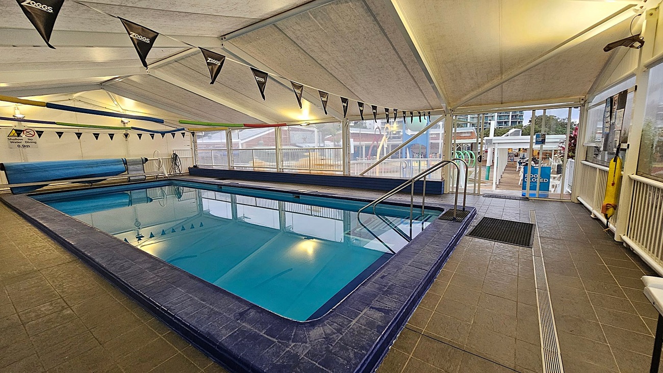 Indoor swimming pool with a blue tiled edge and clear water. Black triangular flags with "Zoggs" hang above. The floor is tiled in grey, and a metal handrail leads into the pool. On the right, a glass door opens to an outdoor area with tables and chairs. The ceiling is white and tent-like, with lights illuminating the space. A sign indicates a shallow water depth of 0.9m.