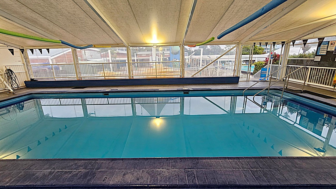 An indoor swimming pool with clear blue water, surrounded by a tiled deck. On the left, there is a hose reel mounted on the wall. The right side features metal handrails leading into the pool and a sign indicating the pool is closed. The ceiling is a white tent-like structure with lights, and the background shows large windows with a view of an outdoor area, including some buildings and greenery.