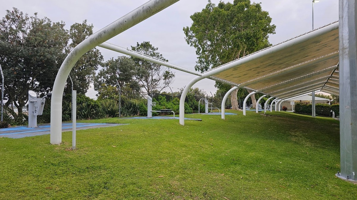 A grassy area with a series of white, curved metal structures forming a canopy on the right. Beneath the canopy, there are exercise machines on blue mats. Trees and bushes line the background, with a fence visible behind them. The ground is covered in well-maintained grass, and the canopy provides shade over the exercise area.