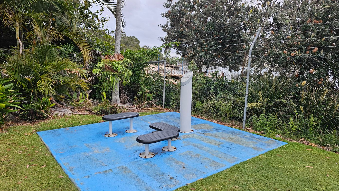 Outdoor exercise area with a blue rubber mat on the ground. Two black, curved benches are mounted on the mat. A tall, white information pillar stands nearby. Surrounding the area are lush green plants and trees, including a palm tree on the left. A chain-link fence with barbed wire runs along the back, with more greenery visible beyond it. The ground around the mat is grassy.