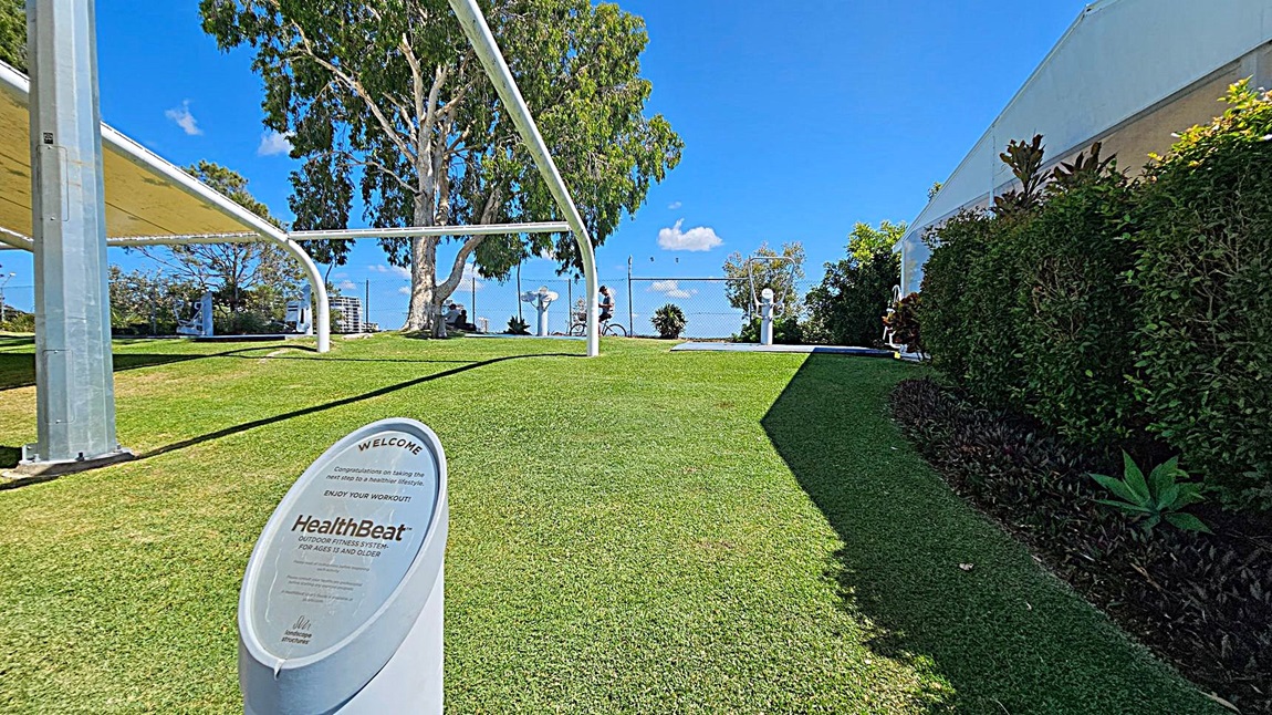 A grassy area with a sign in the foreground that reads "HealthBeat Outdoor Fitness System for Ages 13 and Older." On the left, there are large white metal structures providing shade. In the background, a person is cycling near a fence, and there are trees and shrubs. On the right, a hedge borders the grass.