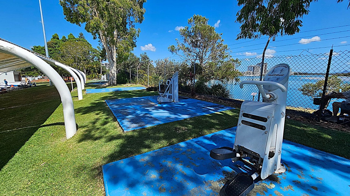 Outdoor gym equipment on blue mats is in the foreground, with a leg press machine visible. To the left, a covered area with white arches and a grassy field. In the background, trees and a chain-link fence separate the area from a body of water, with buildings visible across the water. A person is seated on a bench near the fence. The ground is covered with grass, and the sky is clear.