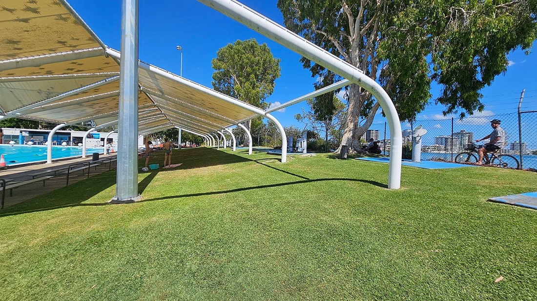A grassy area with a large, white metal canopy structure on the left, providing shade. Underneath, there are benches and a few people standing near a swimming pool. On the right, a person is cycling along a path next to a chain-link fence. Tall trees are scattered throughout, and buildings are visible in the background. The sky is clear and blue.