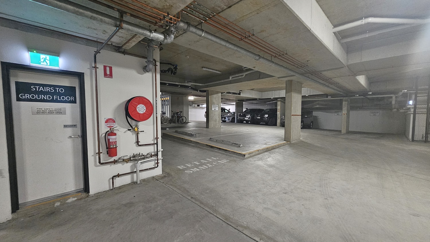 On the left, there is a door labelled "Stairs to Ground Floor" next to a fire hose reel and an extinguisher mounted on the wall. The right side has several parked cars and bicycles near concrete pillars. In the middle, there is a ramp leading to another level of the car park. The background includes more parking spaces and a wall with a sign indicating "Keep Area Clear." The floor is smooth concrete, and the ceiling is concrete with exposed pipes and fluorescent lighting.