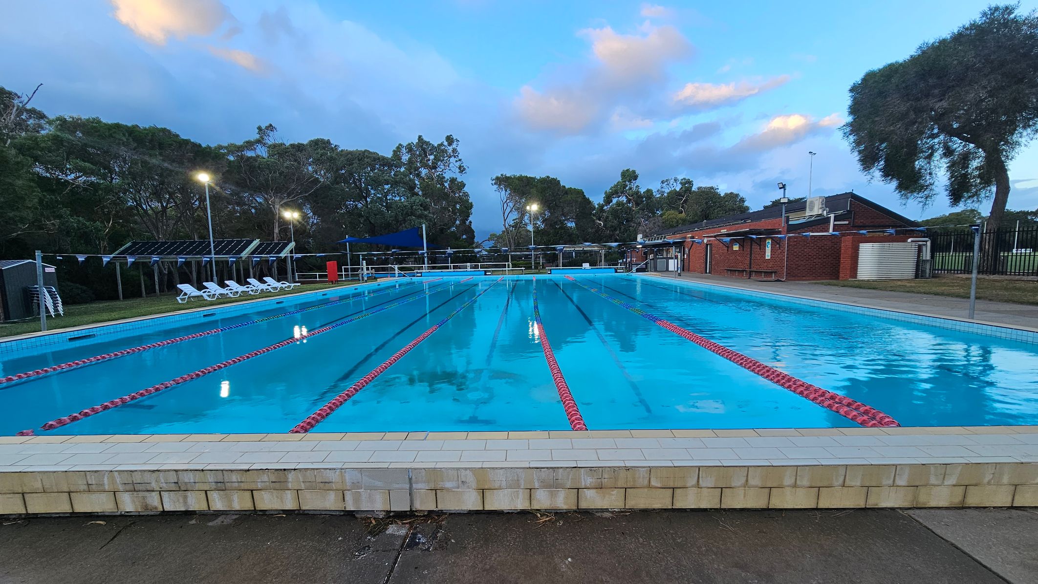 An outdoor swimming pool with clear blue water and red lane dividers stretches into the distance. On the left, there are several white sun loungers under a shaded area. To the right, a brick building with a flat roof is visible, along with a large tree. The background features tall trees and lit lampposts, creating a serene atmosphere. The foreground shows a tiled pool edge and a concrete walkway.