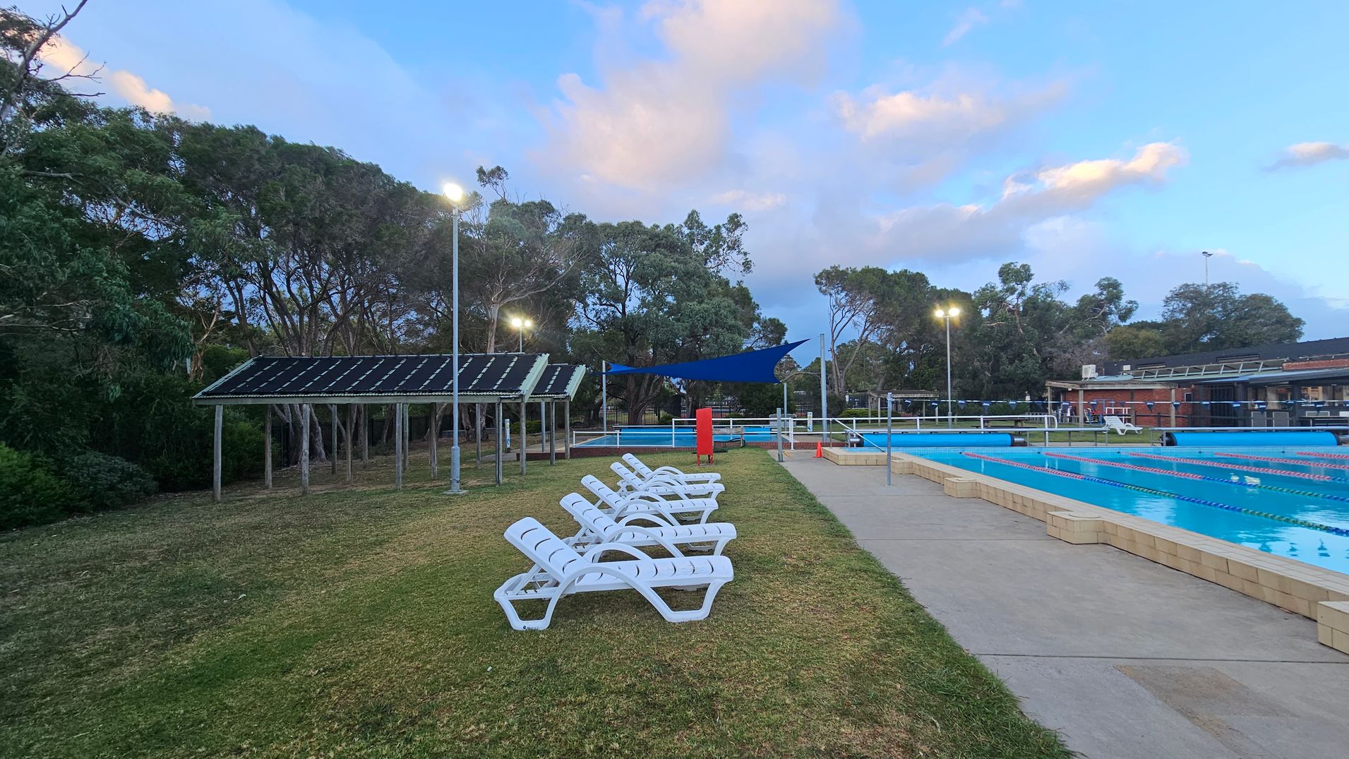 On the left, there are several white sun loungers arranged on a grassy area. Behind them, a structure with a slanted roof is visible, possibly a shelter. In the middle, a blue shade sail is stretched above part of the area. On the right, a swimming pool with lane dividers is bordered by a paved walkway. Trees are in the background, and several tall lights illuminate the scene.