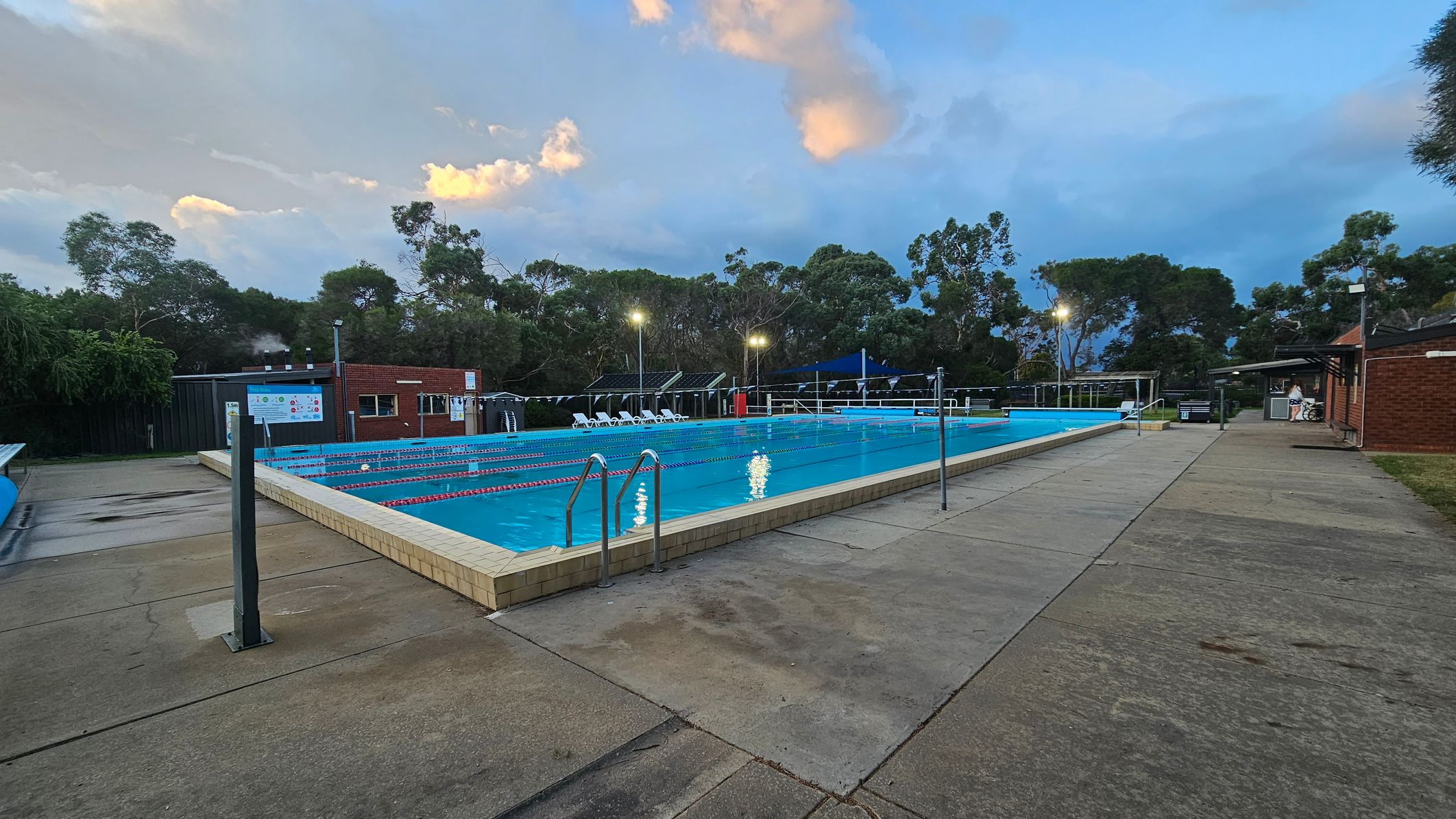 Outdoor swimming pool with lane dividers in the centre, surrounded by a paved area. On the left, there is a signpost and a small building with a noticeboard. On the right, another brick building with a covered area. Trees and shrubs form the background, with some lights illuminating the pool area. The sky is partly cloudy, and the pool water reflects the lights.