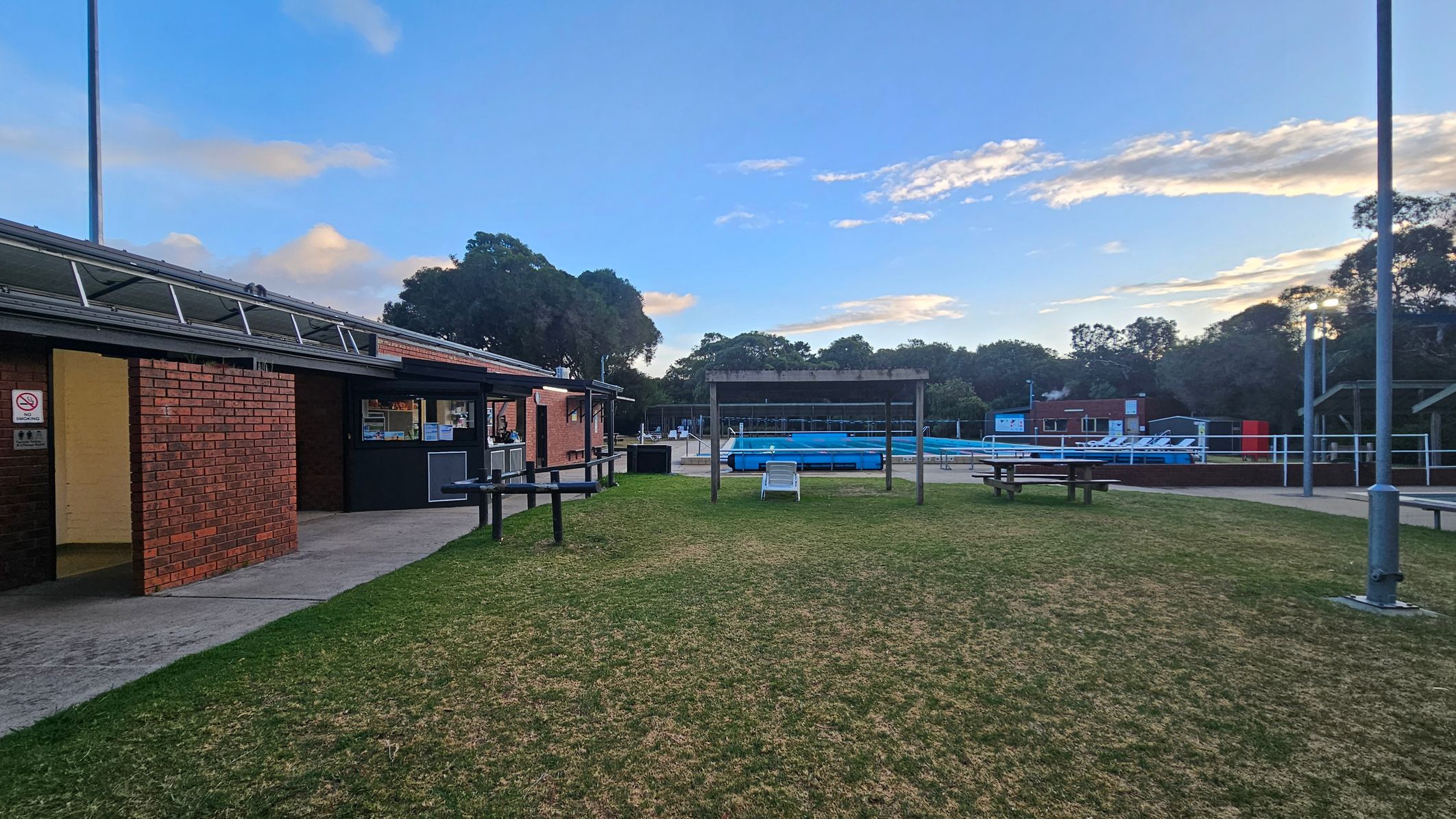 A grassy area with a few picnic tables and a wooden structure in the centre. On the left, there is a brick building with a small service window and an open door. In the background, a swimming pool is visible, surrounded by a fence and some trees. To the right, there are more buildings and a few sun loungers. The area is lit by tall street lamps.