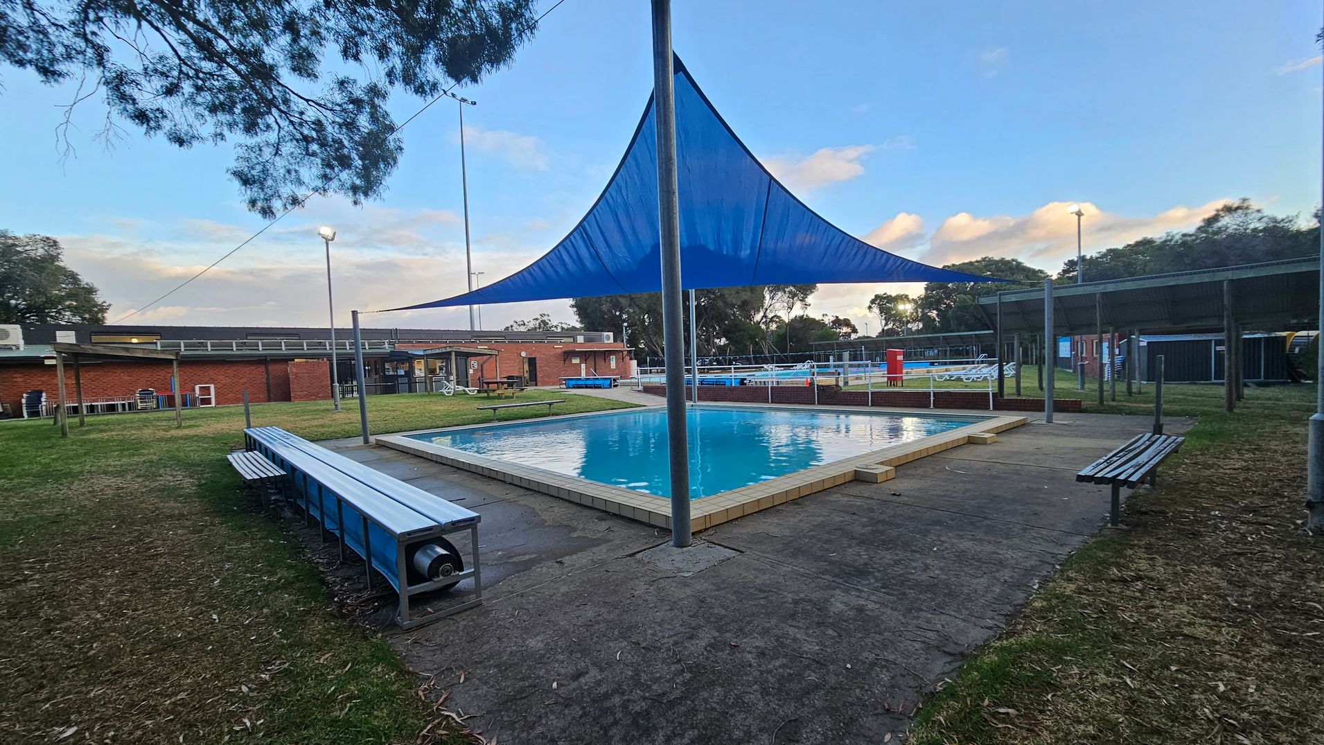 A small outdoor swimming pool is in the centre, surrounded by a paved area. Above the pool, a blue triangular shade sail is supported by a central pole. Benches line the perimeter of the pool area. In the background, there is a brick building with a flat roof and some outdoor seating. Trees and grassy areas are visible around the pool, and there are tall light poles. The sky is partly cloudy.