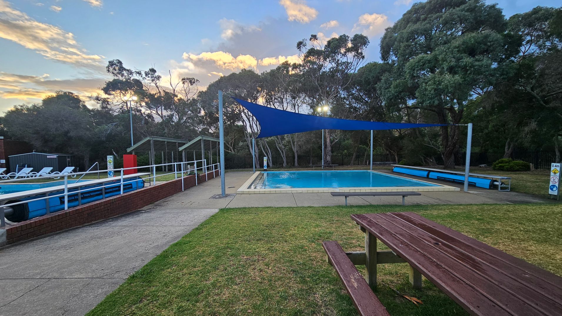 A swimming pool is in the centre, covered by a blue shade sail. On the left, there are white sun loungers and a small building. The pool is surrounded by a low brick wall and a concrete path. On the right, a wooden picnic table sits on a grassy area. In the background, tall trees are visible, and there are two green-roofed shelters. The sky is partly cloudy, and the area is well-lit with outdoor lights.