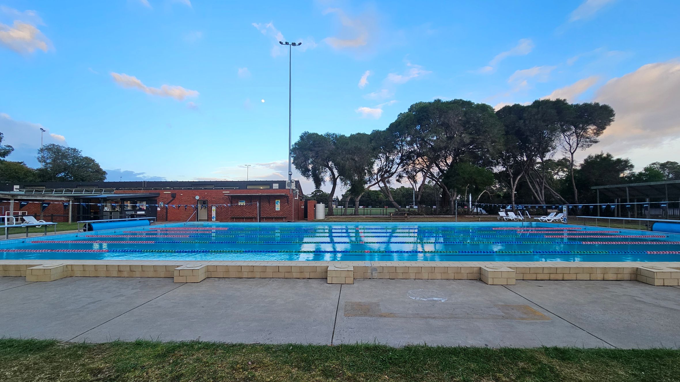 An outdoor swimming pool with lane dividers is in the foreground, surrounded by a paved area and grass. In the background, there is a brick building with a flat roof. To the right, several white sun loungers are visible. Tall trees line the background, and a tall light pole is positioned near the centre. The sky is clear with a few clouds.