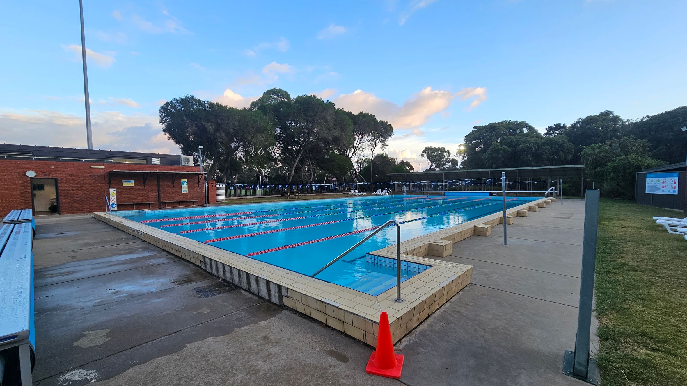 Outdoor swimming pool with clear blue water and red lane dividers in the centre. On the left, a brick building with a door and a long bench. A tall light pole is visible. On the right, a grassy area with white lounge chairs and a fence. A red traffic cone is placed near the pool's edge. In the background, tall trees line the perimeter, and a sign is visible on a fence. The ground is concrete, and the sky is partly cloudy.