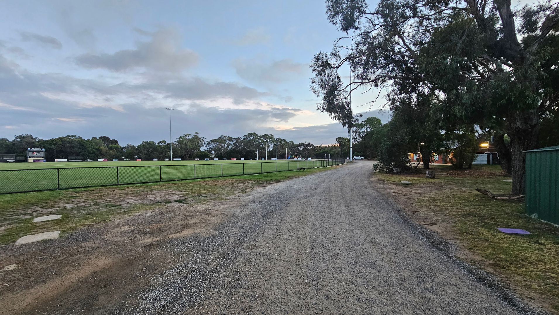 A gravel path leads through an open area. On the left, a white building with a sign featuring a magpie and the words "Home of the Magpies" is visible. The building is surrounded by greenery. On the right, there is a long, low structure with colourful murals and a sign that reads "Casual Court Hire". Tall trees and several lamp posts are in the background.