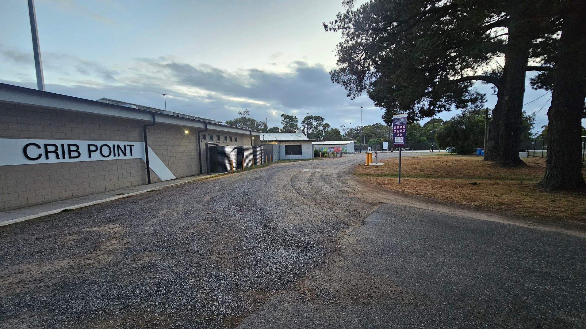 A gravel path curves through an outdoor area. On the left, a building with a sign reading "CRIB POINT" is visible, with several black water tanks lined up against it. On the right, there are large trees and a grassy area with a signpost. In the background, more buildings and trees can be seen, along with a few streetlights. The sky is overcast, casting a muted light over the scene.