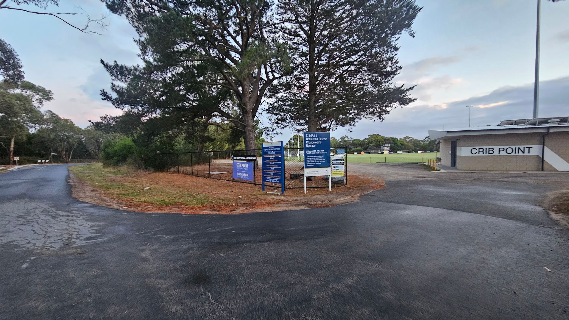 A paved road curves to the left, bordered by trees and grass. In the centre, there are several signs near a large tree, with a fence behind them. On the right, a building with "CRIB POINT" written on it is visible, adjacent to a sports field with goalposts. The ground is asphalt, and the area is surrounded by greenery.