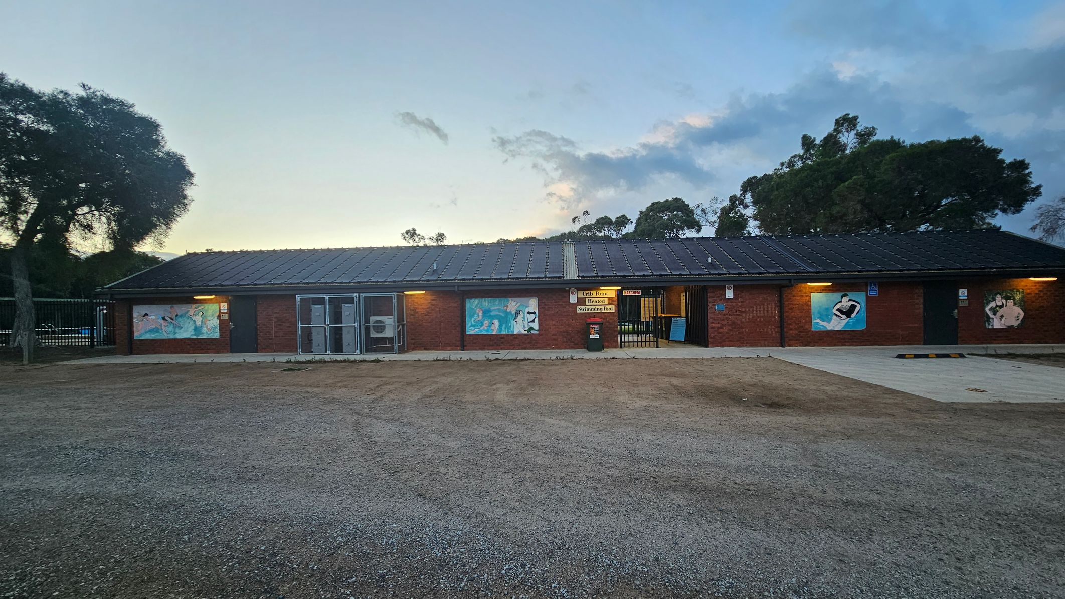 A single-storey brick building with a dark roof, featuring three large murals on its exterior walls. The entrance is centrally located, with a sign above it. The ground in front is a mix of gravel and sparse grass. Trees are visible in the background, and the sky is overcast. The building is lit by exterior lights, casting a warm glow on the walls.