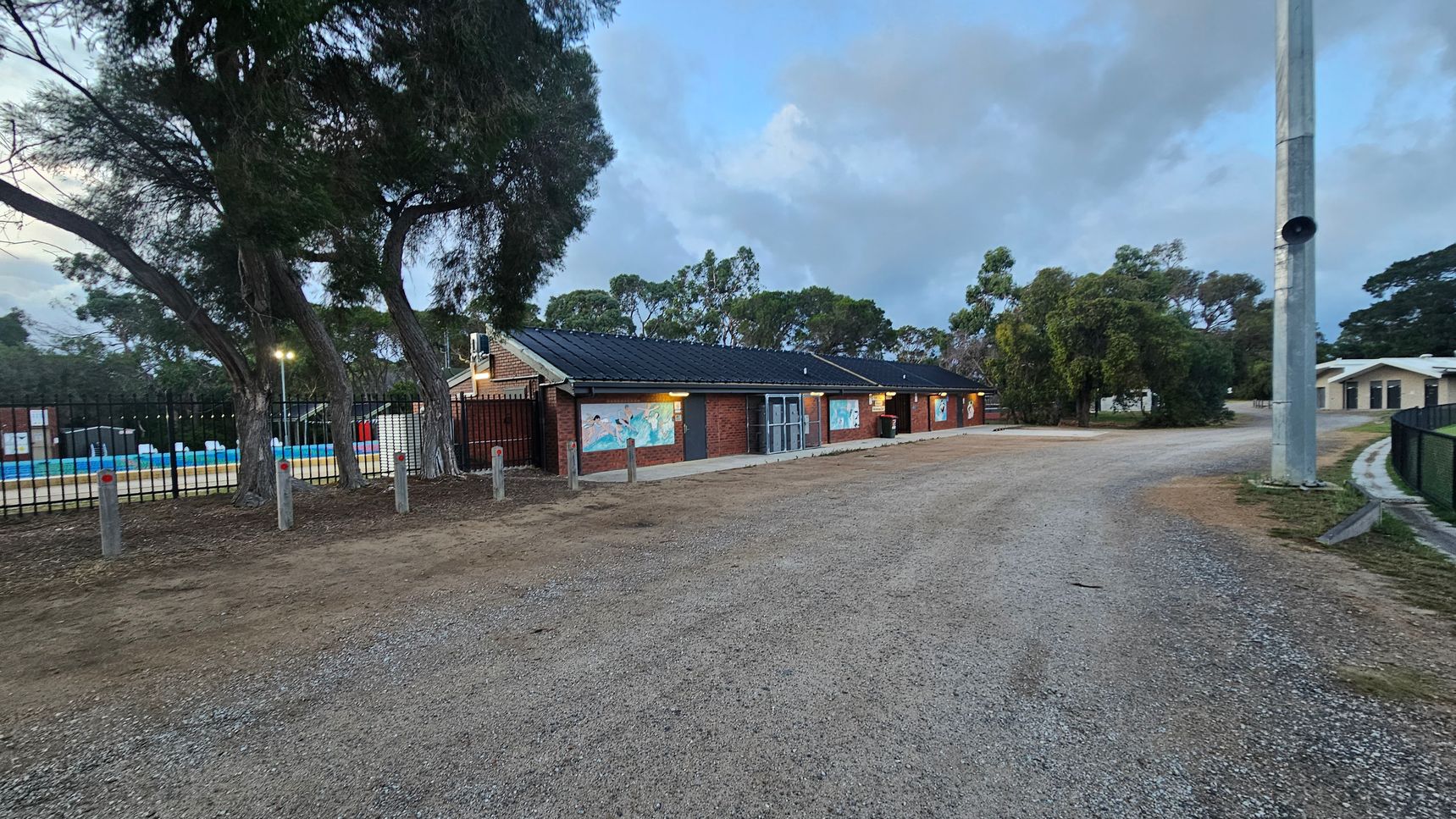 A gravel path leads to a brick building with a dark roof in the centre. The building has murals on its walls and is surrounded by trees. On the left, a fenced area with a pool is visible, with tall trees nearby. On the right, a tall pole stands next to a grassy area, and another building is partially visible in the background. The sky is overcast, and the area is well-lit with outdoor lights.