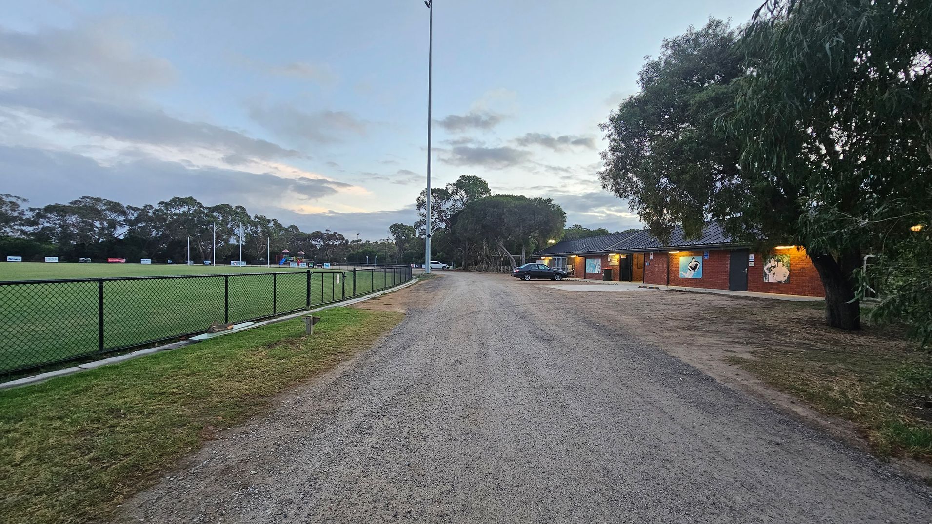 A gravel path runs through the centre, with a sports field on the left, enclosed by a chain-link fence. The field has several goalposts and advertising boards. On the right, there is a brick building with illuminated signs and a parked car nearby. Trees are scattered around the area, with some overhanging the building. The sky is overcast, and the ground is a mix of grass and gravel.