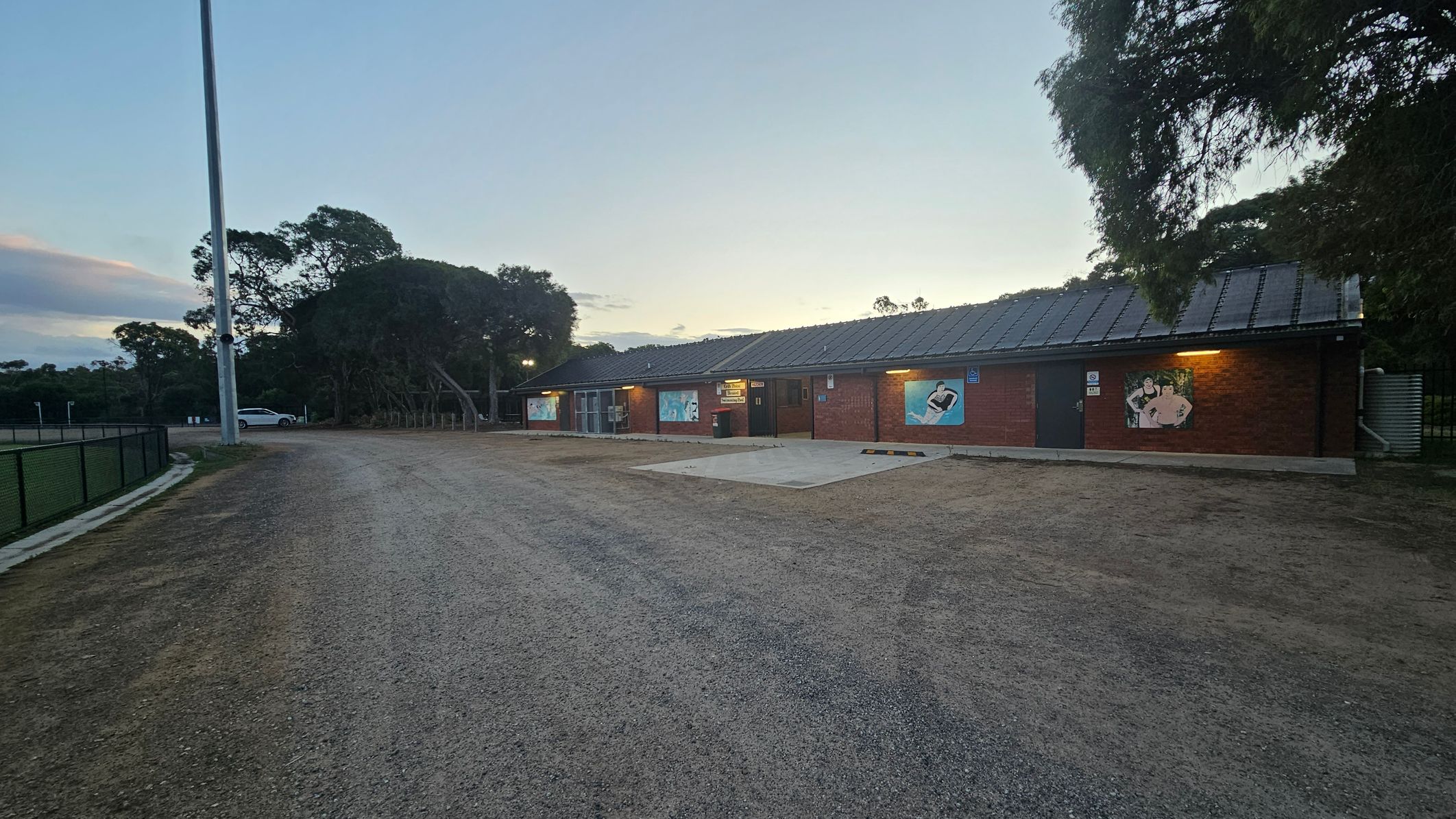 A single-storey brick building with a dark roof is on the right, featuring several large painted murals on its walls. The building is illuminated by exterior lights. On the left, a gravel path runs alongside a fenced area, with a tall light pole and a parked white car visible. Trees are in the background, and the sky is a gradient of light colours, suggesting early morning or late evening. The ground is a mix of gravel and patches of grass.