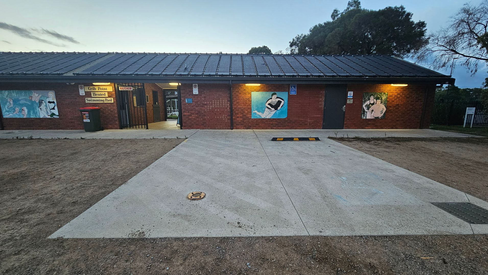 A brick building with a dark roof, featuring three murals on the front wall. On the left, a mural with multiple figures, and a sign above reading "Crib Point Heated Swimming Pool." A red bin is nearby. In the centre, a mural of a person in water. On the right, another mural with two figures. The ground is a mix of concrete and dirt, with a small ramp and a drain visible. Trees are in the background.