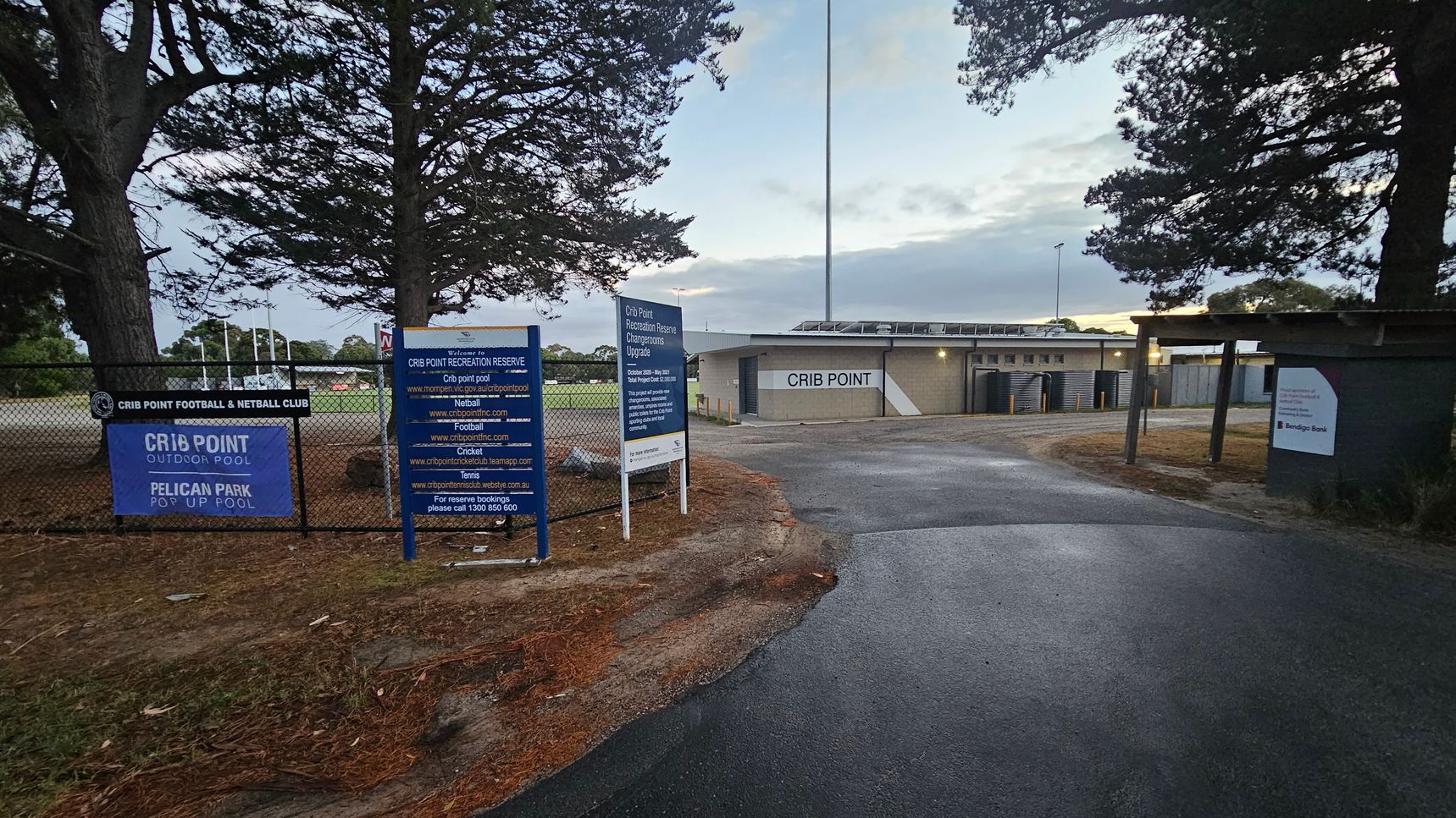 On the left, there are several signs on a chain-link fence, including ones for Crib Point Football & Netball Club and Crib Point Outdoor Pool. Behind the fence, trees and a grassy area are visible. On the right, a paved path leads to a building with "Crib Point" written on it. The building has a flat roof and is surrounded by more trees. The ground is a mix of asphalt and dirt, and there are tall light poles in the background.
