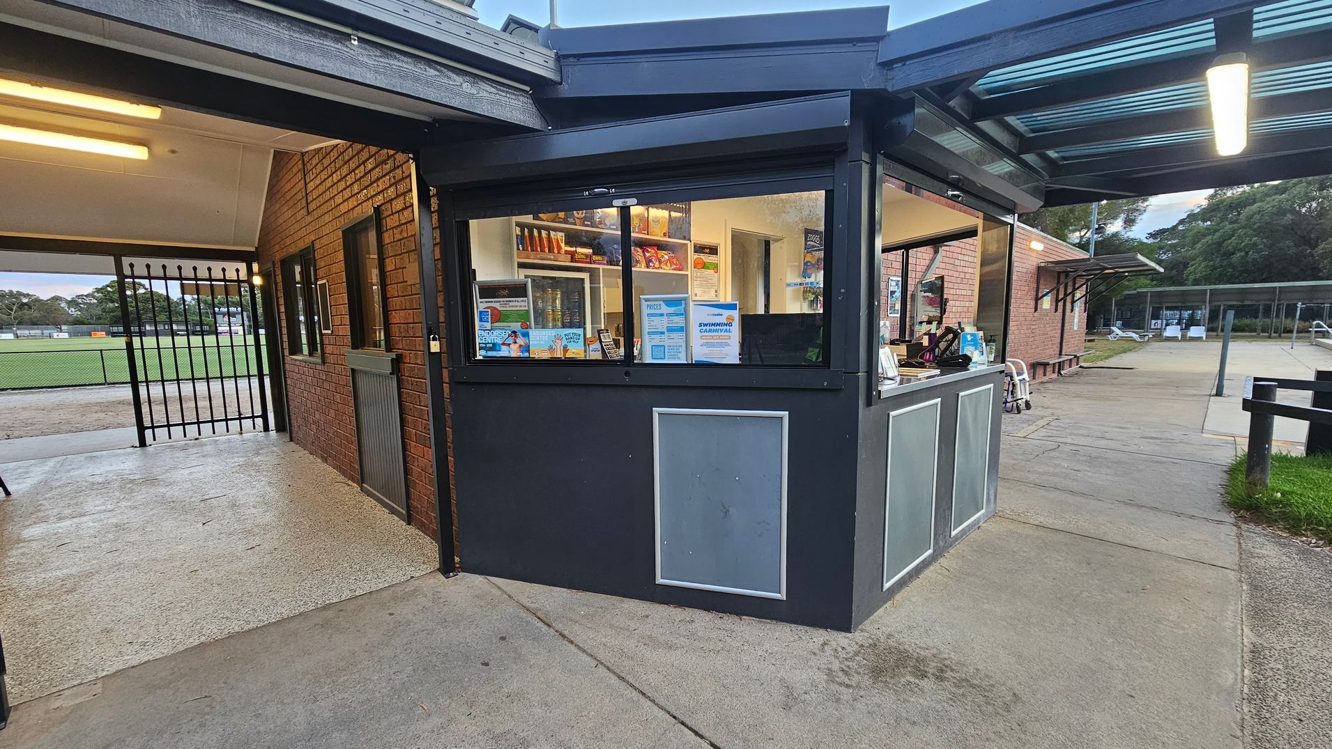 A brick building with a metal roof. In the centre, a gated entrance leads to a corridor with a sign reading "HAZCHEM" above. On the left, a sign says "Crib Point Heated Swimming Pool" and a rubbish bin with a red lid is nearby. A mural of a swimmer is painted on the wall. On the right, another mural of a swimmer is visible. The floor is concrete, and there are trees in the background.