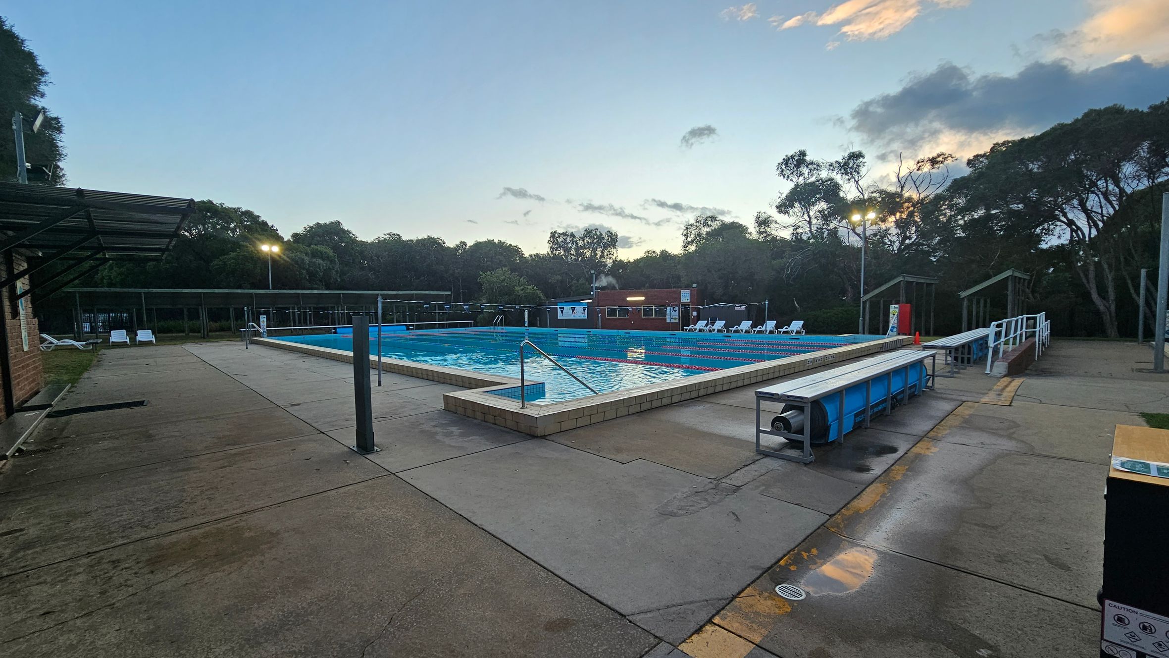 Outdoor swimming pool surrounded by a concrete deck. On the left, there is a covered area with a few white sun loungers. The pool is in the centre, with lane dividers visible. On the right, there are more sun loungers and a small building. Trees line the background, and there are a few lights illuminating the area. The foreground shows a pool cover roller and a caution sign. The sky is partly cloudy.