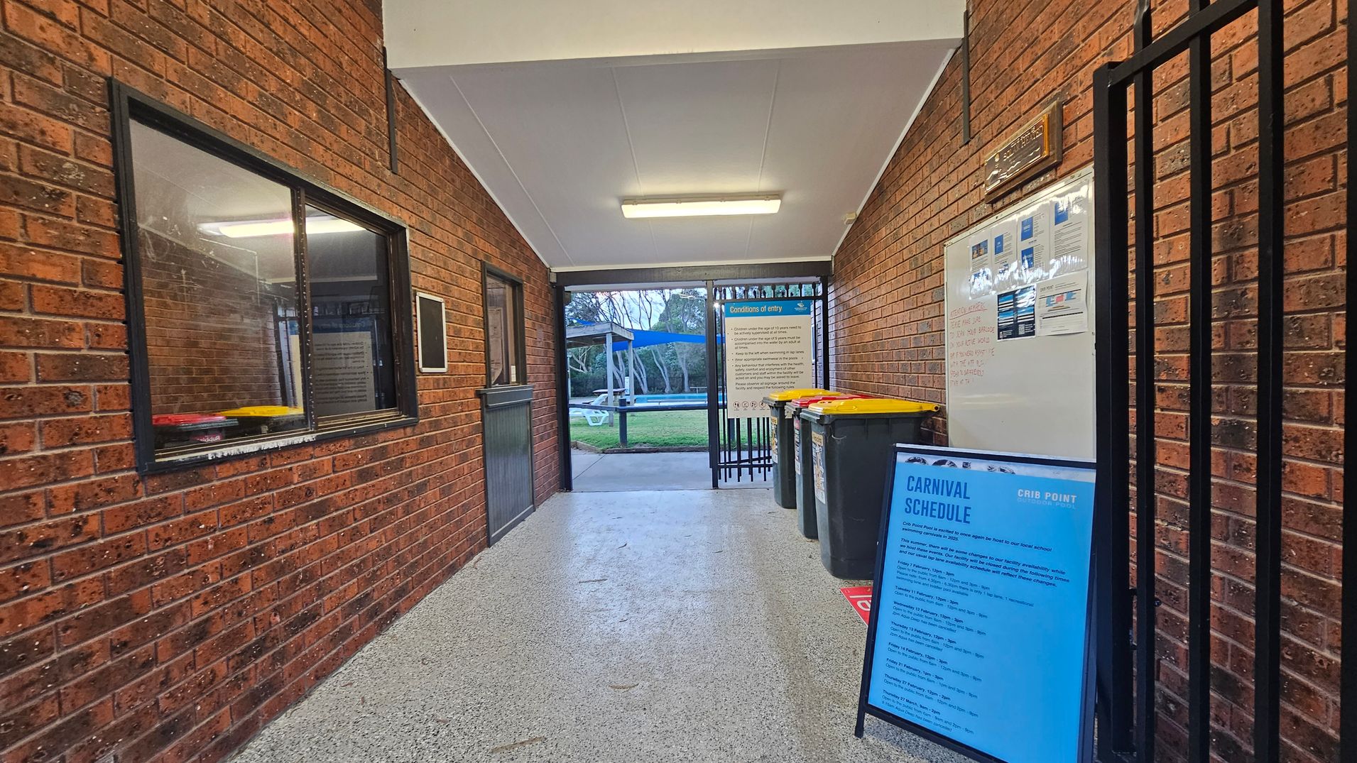 A corridor with brick walls on both sides. On the left, there's a window with a black frame and a small ledge. On the right, a whiteboard with notices and a blue sign titled "Carnival Schedule" is visible. Several bins with yellow lids are lined up against the right wall. The floor is speckled grey, and the ceiling is white with a fluorescent light fixture. At the end of the corridor, there's an open gate leading to a grassy area with trees and a blue shade structure.