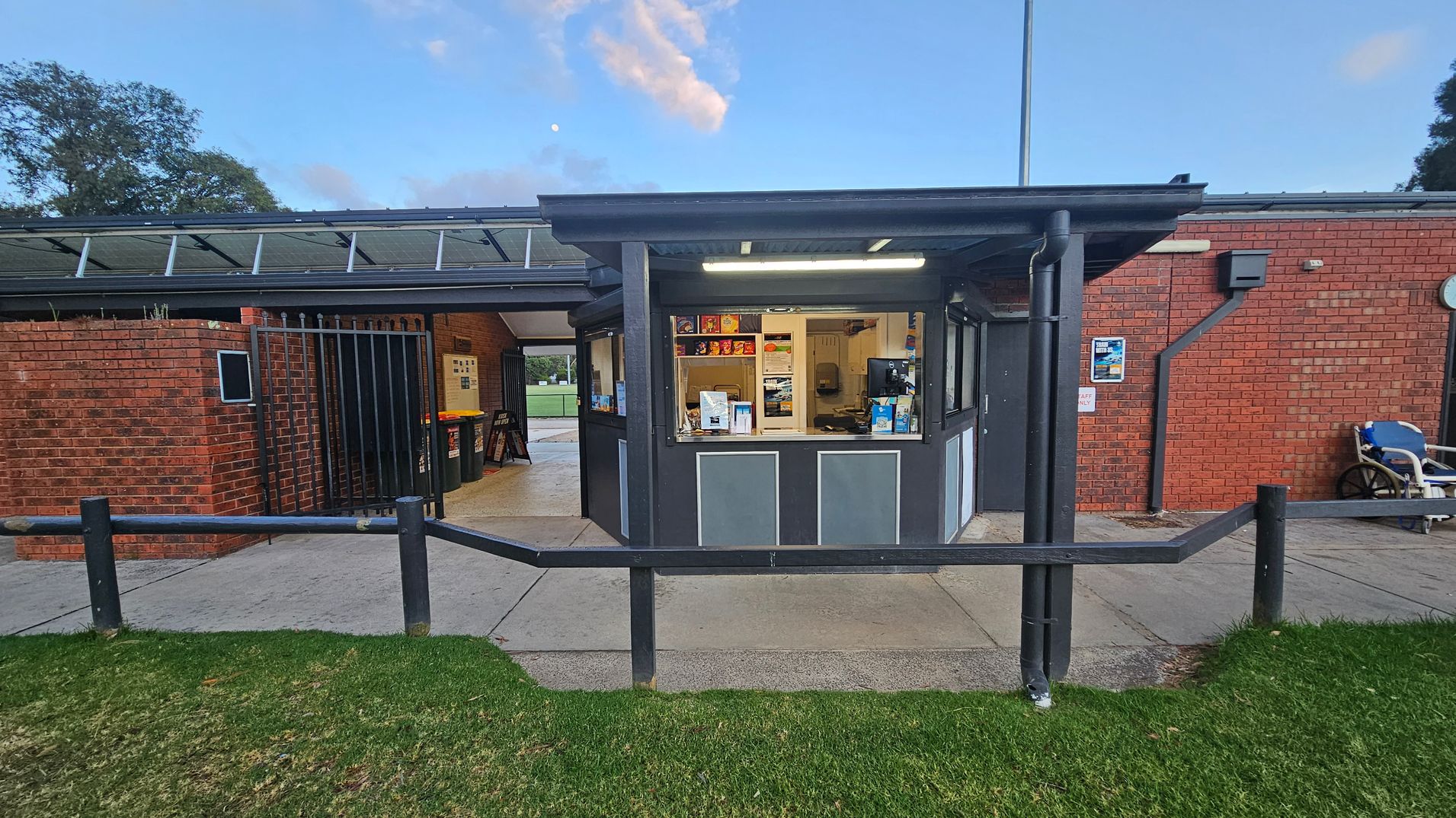 A small kiosk with a serving window is in the centre, displaying snacks and drinks. The structure is made of brick and metal, with a flat roof. To the left, there is a brick wall with a gated entrance leading to an interior space. On the right, a brick wall features a poster and a wheelchair parked nearby. The ground is a mix of concrete and grass, and a wooden railing runs across the foreground. Trees are visible in the background.
