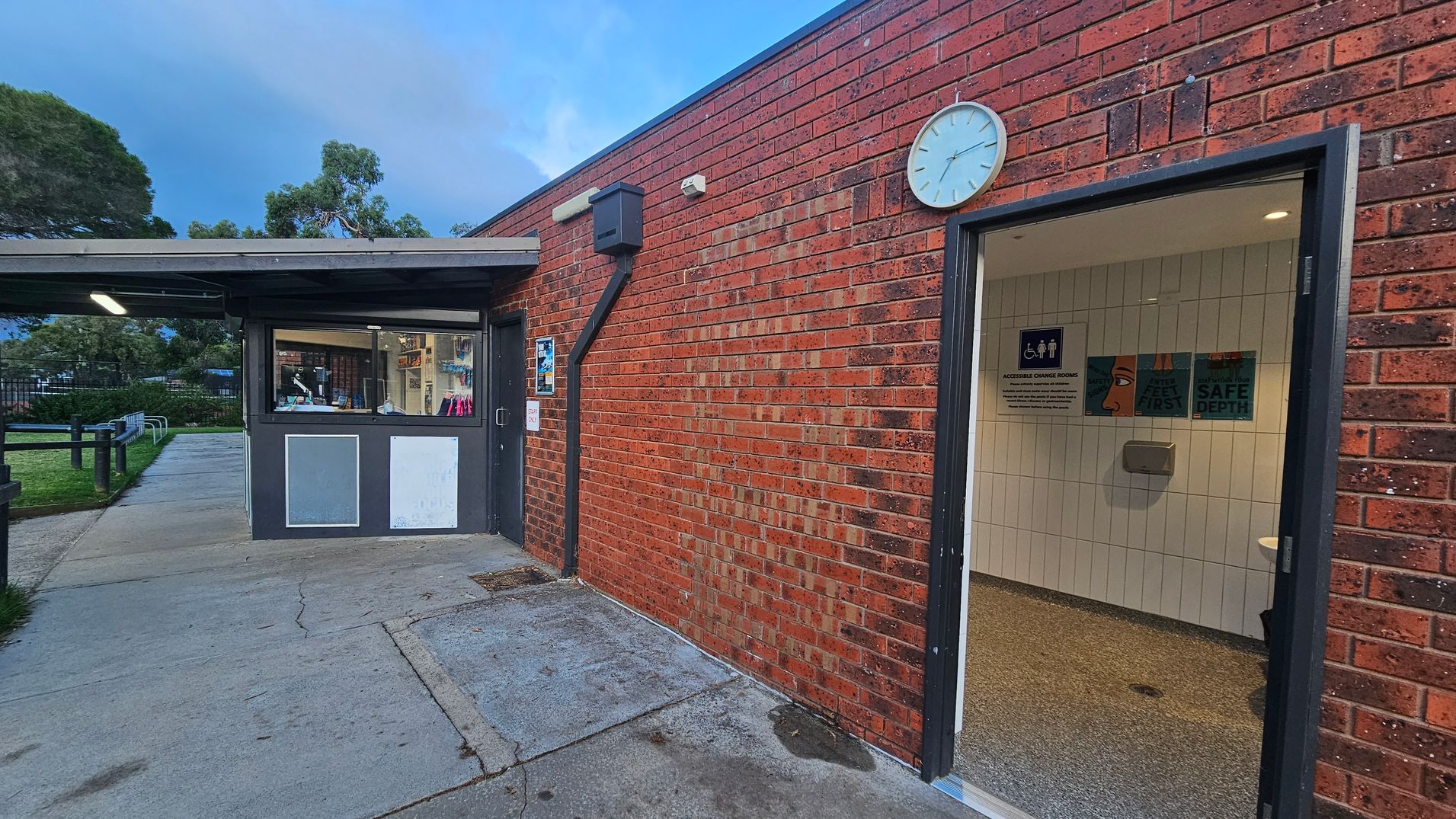 On the left, there is a building with a window displaying various items inside, and a pathway leading to a grassy area with trees. On the right, a brick wall features a clock and an open door leading to a tiled room with safety signs. The floor is concrete outside and tiled inside the room. The ceiling is not visible.