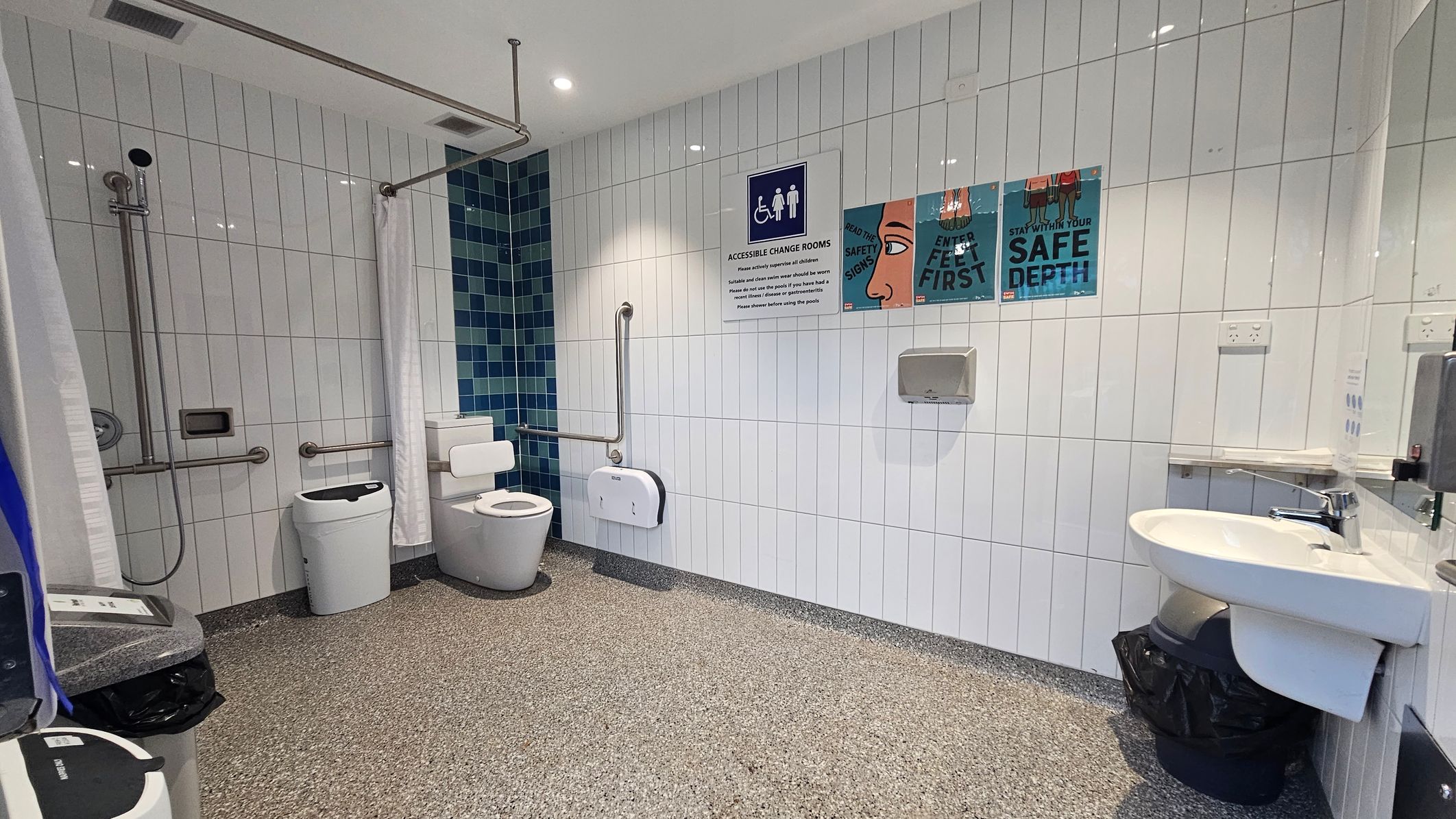 Accessible bathroom with white tiled walls and a speckled floor. On the left, a shower area with a curtain, grab bars, and a showerhead. Next to it, a toilet with support rails and a nappy bin. The right side features a sink with a mirror above and a soap dispenser. The wall displays signs about safety and accessibility. The ceiling is white with recessed lighting.